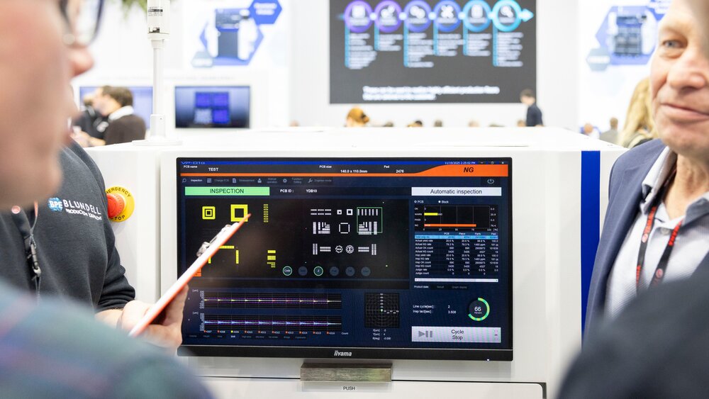 Mehrere Personen stehen gemeinsam um den Monitor einer Maschine herum, die an einem Stand auf der productronica ausgestellt wird.