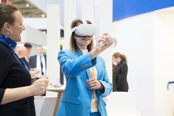 Eine Besucherin auf der transport logistic, die ein VR-Headset und einen Controller verwendet, während eine Ausstellerin neben ihr steht.
