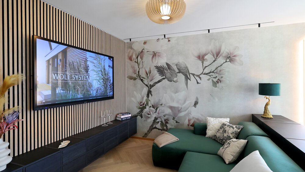 Ein moderner Wohnbereich mit senkrechten Holzlatten an der Fernsehwand und einer Vogeltapete daneben. Vor dem Fernseher steht eine grüne Couch.