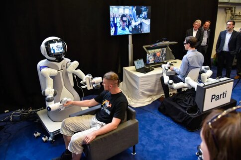 Eine Person interagiert mittles Telepräsenz über einen humanoiden Roboter mit einer zweiten, sitzenden Person im selben Raum.