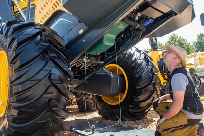 Mann begutachtet die Unterseite eines großen Forst- oder Landfahrzeugs mit massiven Reifen auf dem Messegelände.