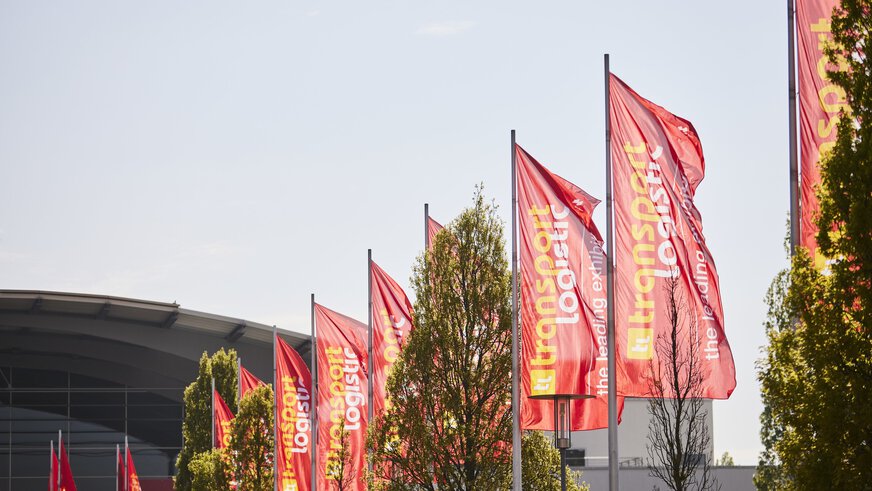 Viele rote Fahnen mit dem Logo der transport logistic wehen vor dem Eingang zur Messe München.