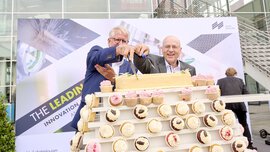 Zwei Männer schneiden vor einer Messehalle eine große, längliche Torte auf einem Etagere mit Cupcakes an.