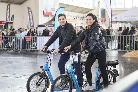 Ein Testbereich für Fahrräder, in dem ein Mann und eine Frau je ein blaues Fahrrad zur Probe fahren, während im Hintergrund Messebesucher hinter dem Zaun stehen.