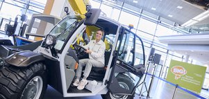 Young woman sitting at the wheel of a telescopic handler in front of Think Big sign.