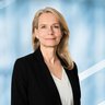 Eine Frau mit langen blonden Haaren und blauen Ohrringen, in einer weißen Bluse mit einem schwarzen Jackett, lächelt.