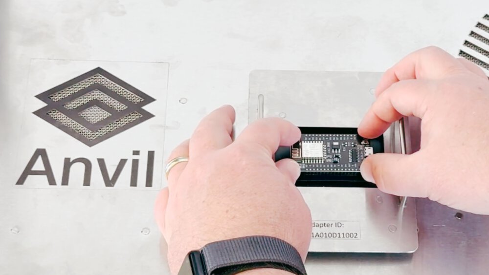 Two hands fit a circuit board onto a metal base. Next to it is the angular logo of “Anvil” on a metal surface.