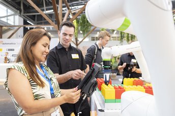 Menschen interagieren an einem Messestand mit einem Roboterarm, der bunte Steckbausteine platziert.