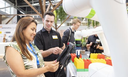 Menschen interagieren an einem Messestand mit einem Roboterarm, der bunte Steckbausteine platziert.