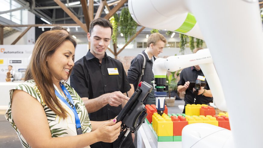 People interacting at a trade fair stand with a robotic arm that places colorful building blocks.