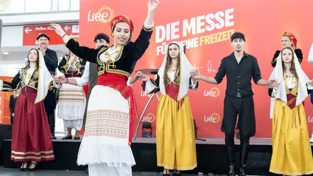 A folklore group in traditional costumes performs a dance on a stage in front of the trade fair logo, while other dancers stand in the background.
