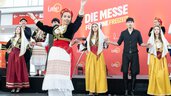 A folklore group in traditional costumes performs a dance on a stage in front of the trade fair logo, while other dancers stand in the background.