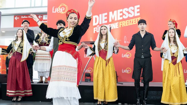 A folklore group in traditional costumes performs a dance on a stage in front of the trade fair logo, while other dancers stand in the background.