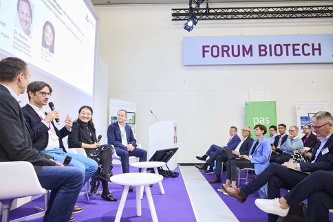 Four people sit in armchairs and talk to each other. One of the people is holding a microphone and gesticulating while talking
