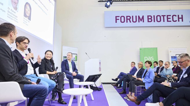 Four people sit in armchairs and talk to each other. One of the people is holding a microphone and gesticulating while talking