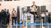 Girl jumps over a hobby-horse hurdle inside an exhibition hall, onlookers stand nearby, another child waits in the background; a lively, energetic moment captured.
