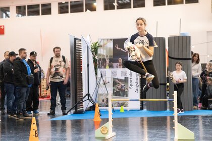 Mädchen springt mit Steckenpferd über Hindernis in einer Messehalle, Zuschauer knapp im Hintergrund, weiteres Kind wartet bereit, dynamische Szene.