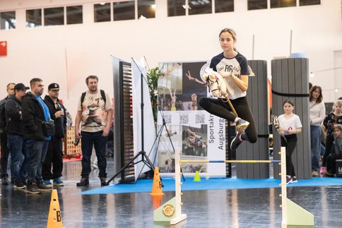 Mädchen springt mit Steckenpferd über Hindernis in einer Messehalle, Zuschauer knapp im Hintergrund, weiteres Kind wartet bereit, dynamische Szene.