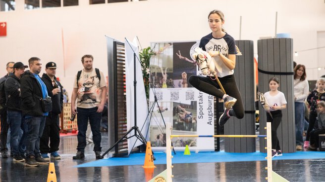 Girl jumps over a hobby-horse hurdle inside an exhibition hall, onlookers stand nearby, another child waits in the background; a lively, energetic moment captured.