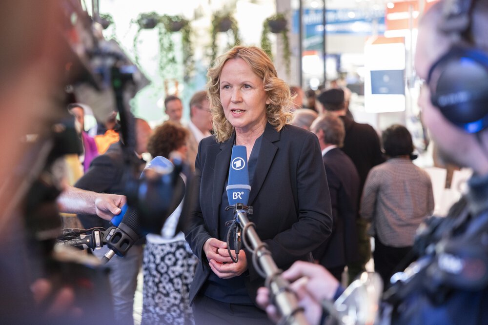A woman in a suit gives a television interview in front of a trade fair stand.