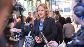 A woman in a suit gives a television interview in front of a trade fair stand.