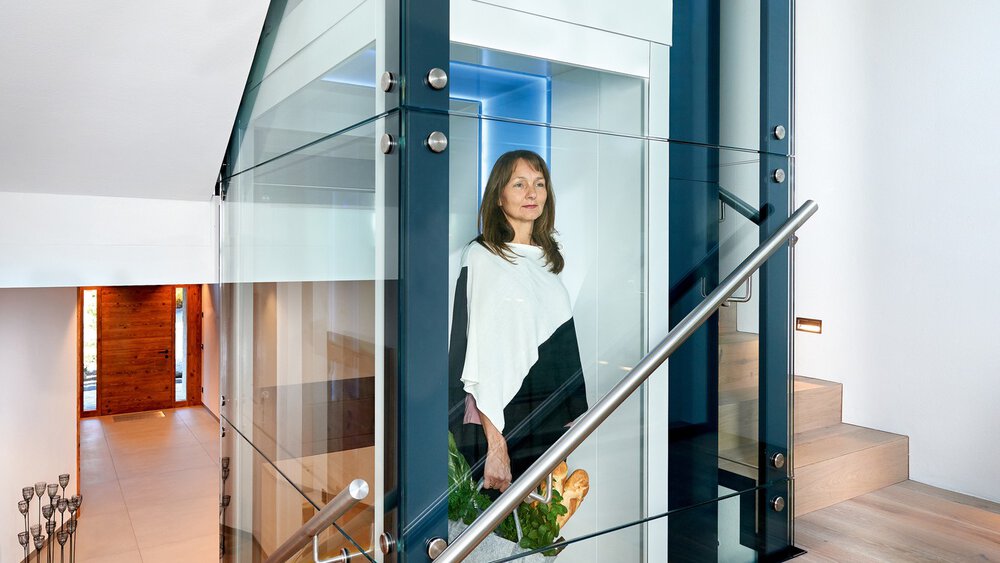 Frau in einem modernen Glasaufzug neben einer Treppe in einem hellen, zeitgenössischen Wohninterieur.