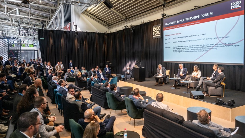 Eine Podiumsdiskussion auf einem Wirtschaftsforum mit einem sitzenden Publikum und einem Präsentationsbildschirm im Hintergrund.