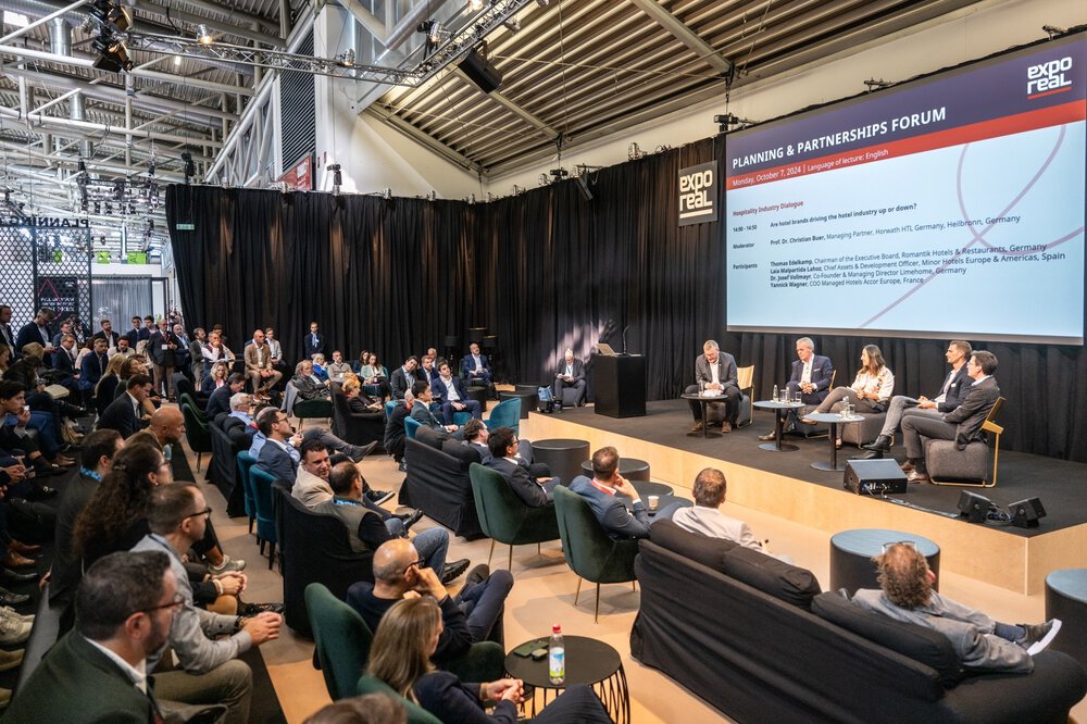 Eine Podiumsdiskussion auf einem Wirtschaftsforum mit einem sitzenden Publikum und einem Präsentationsbildschirm im Hintergrund.