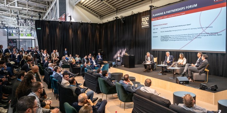 Eine Podiumsdiskussion auf einem Wirtschaftsforum mit einem sitzenden Publikum und einem Präsentationsbildschirm im Hintergrund.