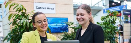 Zwei Frauen im Business-Outfit stehen in der „Career Area“ einer Messe, lächeln und besprechen etwas am Laptop, umgeben von Pflanzen und Messeständen.