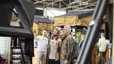 Three people look fascinated at a roof tent at an exhibition stand.