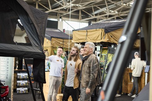 Drei Personen schauen sich fasziniert ein Dachzelt an einem Messestand an.
