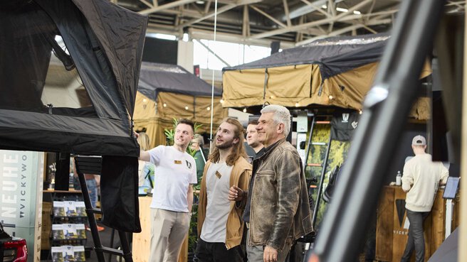 Drei Personen schauen sich fasziniert ein Dachzelt an einem Messestand an.