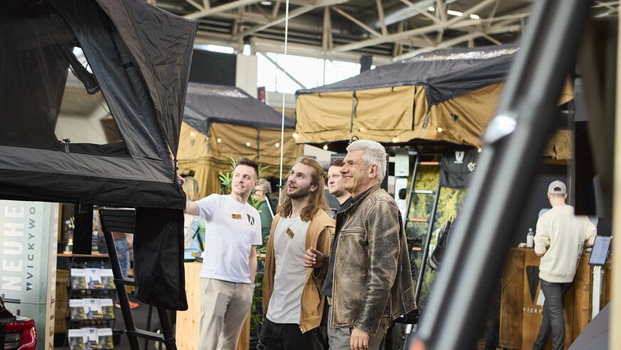 Three people look fascinated at a roof tent at an exhibition stand.
