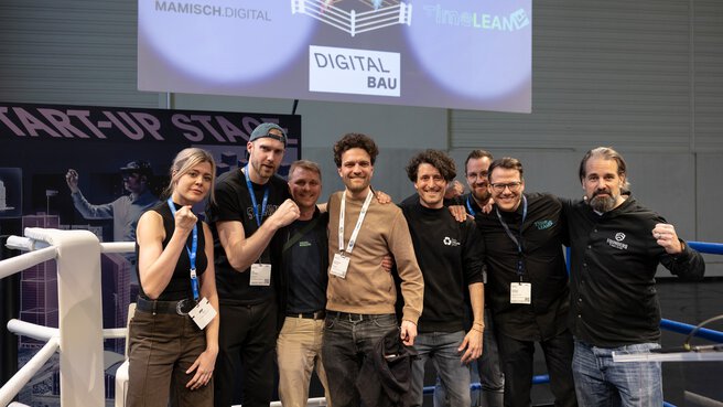 Eine Gruppe von 8 Personen steht in einem Boxring, einige zeigen humorvoll ihre Muskeln