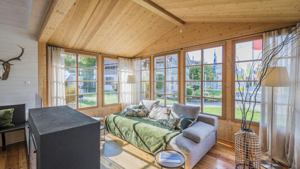 Gemütlicher Wintergarten mit großen Fenstern, einem grünen Sofa, einem Fernseher und einem an der Wand hängenden Hirschkopf.