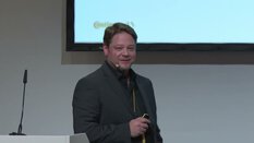 Ein Mann im dunklen Anzug mit Headset hält einen kleinen Clicker und spricht vor einer Leinwand während einer Präsentation.