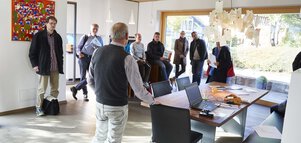 Eine Gruppe von Menschen steht im Wohnzimmer eines Musterhauses und hört aufmerksam einem Redner bei einer Führung zu..