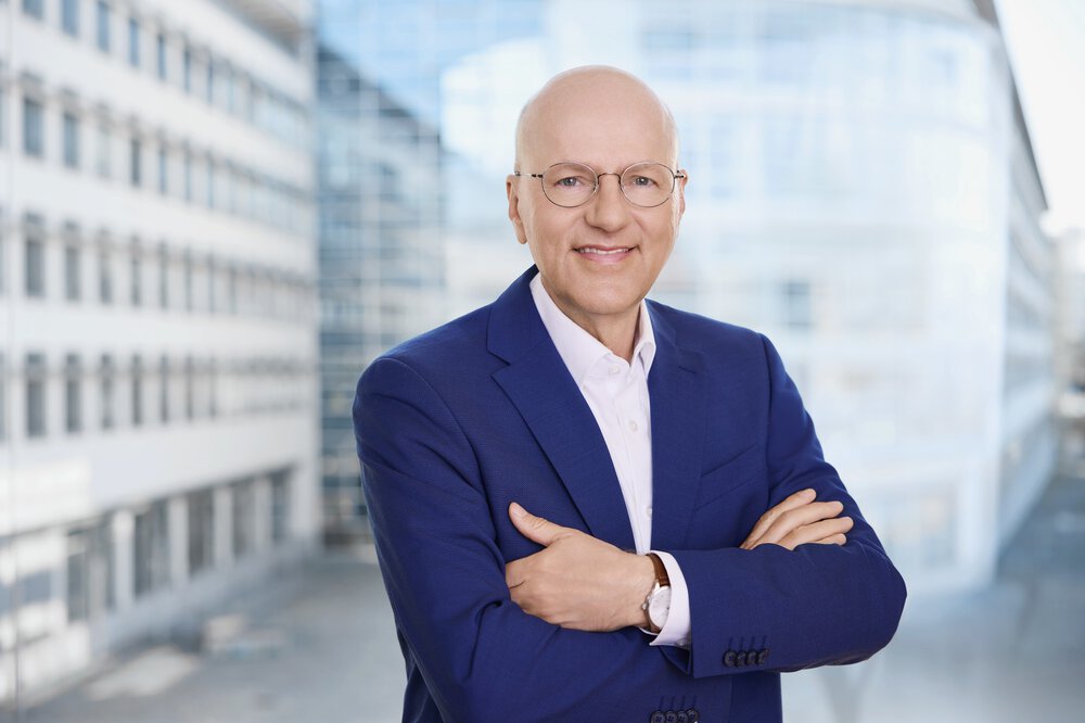 Ein glatzköpfiger Mann mit Brille und blauer Anzugsjacke steht lächelnd mit verschränkten Armen vor einem modernen Gebäude.