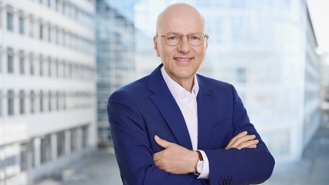 Ein glatzköpfiger Mann mit Brille und blauer Anzugsjacke steht lächelnd mit verschränkten Armen vor einem modernen Gebäude.