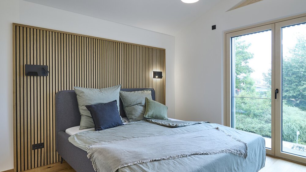 Ein gemütliches Schlafzimmer mit einem ordentlich gemachten Bett, Lampen und einem großen Fenster mit Blick ins Grüne draußen.