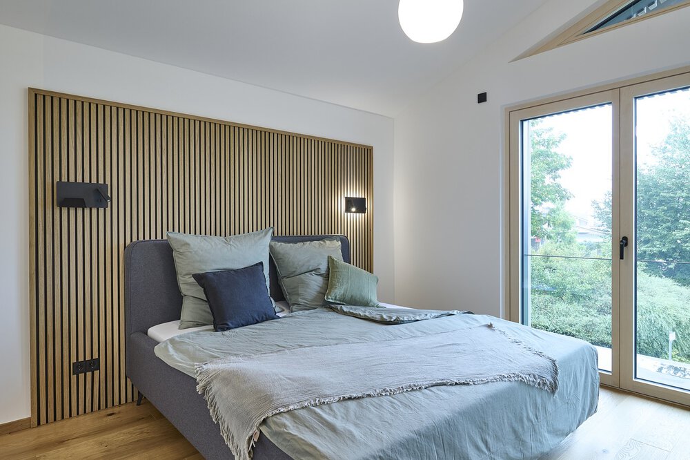 Ein gemütliches Schlafzimmer mit einem ordentlich gemachten Bett, Lampen und einem großen Fenster mit Blick ins Grüne draußen.