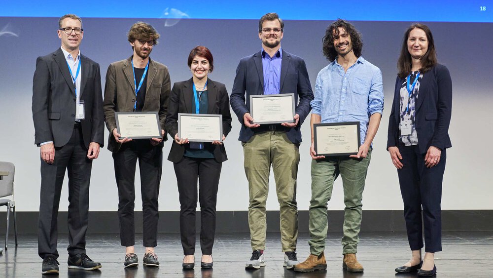 Six people are smiling on stage, four of them holding a certificate in their hands.