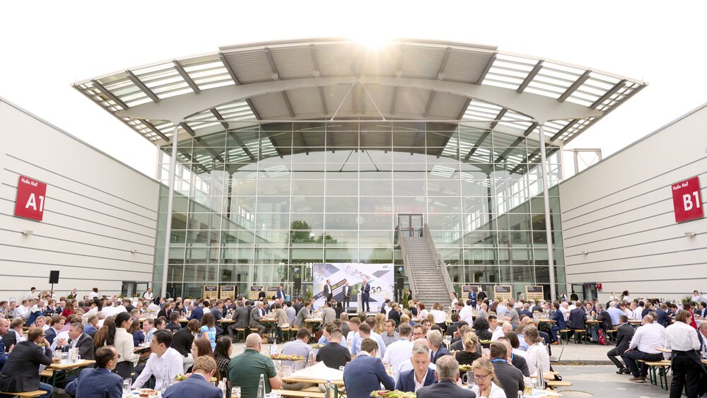Zahlreiche Menschen essen an Biergarnituren auf dem Freigelände zwischen den Messehallen A1 und B1. Im Hintergrund ist eine Bühne zu sehen.