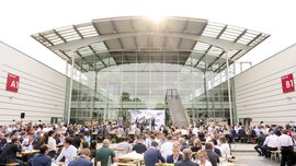 Zahlreiche Menschen essen an Biergarnituren auf dem Freigelände zwischen den Messehallen A1 und B1. Im Hintergrund ist eine Bühne zu sehen.