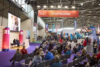 Links ist eine Bühne, auf der 2 Personen ein Gespräch führen. Rechts sitz das Publikum. Oben ist ein Schild mit der Aufschrift Sportbühne. Im Hintergrund sind mehrere SUP-Boards und Sportequipment.