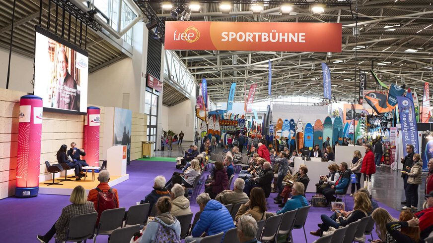 On the left is a stage on which 2 people are having a conversation. The audience is sitting on the right. At the top is a sign saying Sportbühne. In the background are several SUP boards and sports equipment.