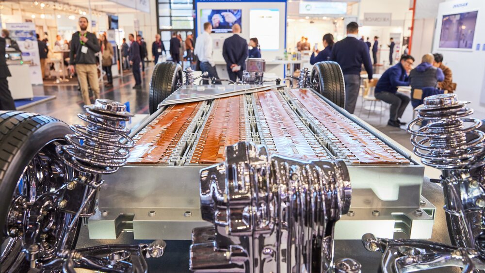 A chassis of an electric car with a large battery pack is on display at a trade fair stand; visitors and other stands can be seen in the background.