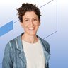 Eine lächelnde Frau mit dunklen kurzen gelockten Haaren trägt Ohrringe, ein weißes Shirt und eine blaue Strickjacke darüber.