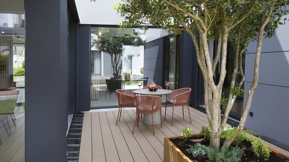 Moderne Terrasse mit rundem Tisch, braunen Stühlen und großem Topfbaum, umgeben von dunklen Wänden und Glasfenstern.